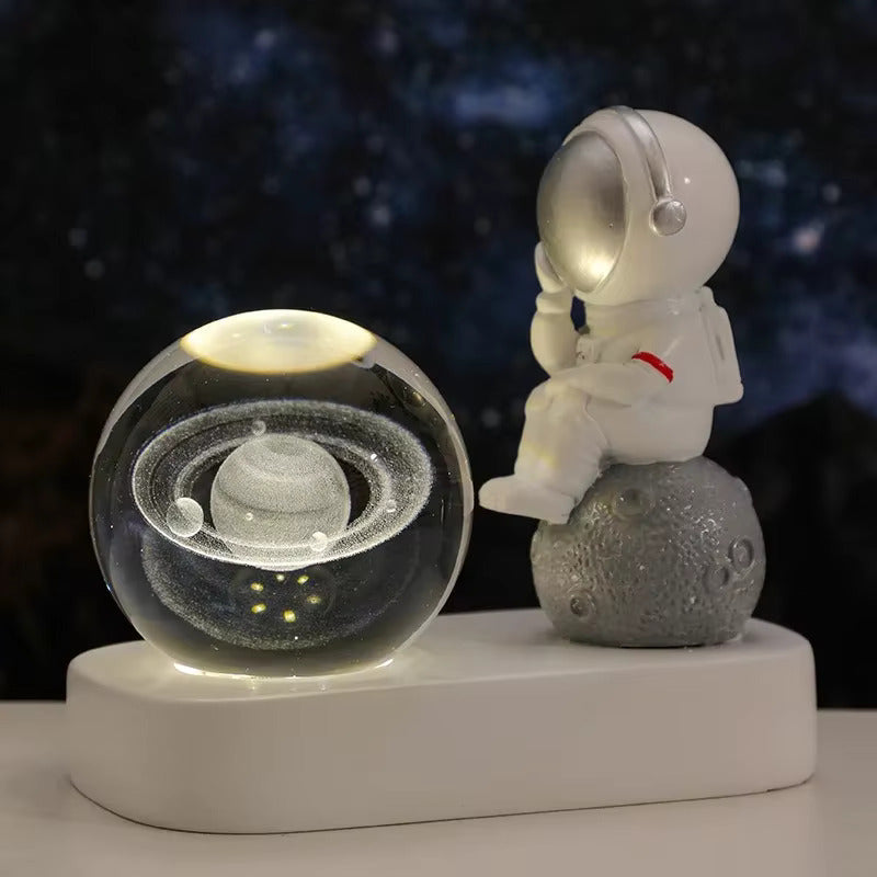 Luminária decorativa com astronauta sentado sobre a lua, observando uma esfera iluminada que exibe o planeta Saturno com seus anéis, contra um fundo estrelado.