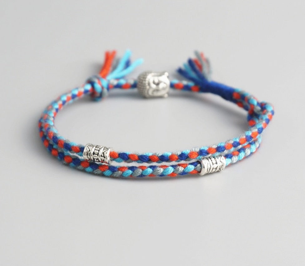 Pulseira Budista trançada em fios azul e laranja, com detalhes prateados de Buda e símbolos espirituais. Pelicano Azul.