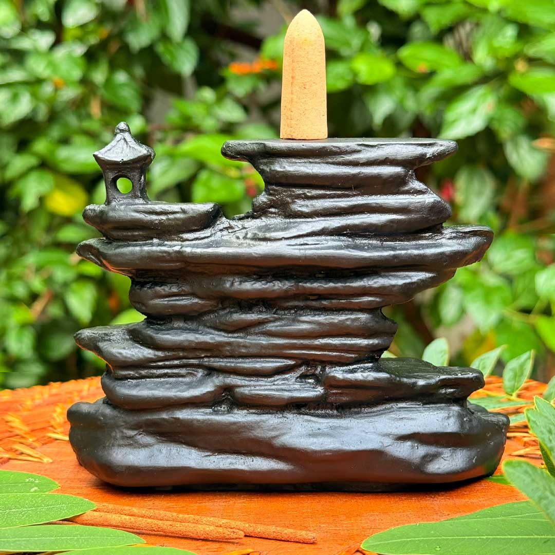 Incensário Cachoeira de Rozan Pelicano Azul em preto, com formato de rocha esculpida e pequeno pagode, posicionado sobre superfície natural decorada com folhas verdes.