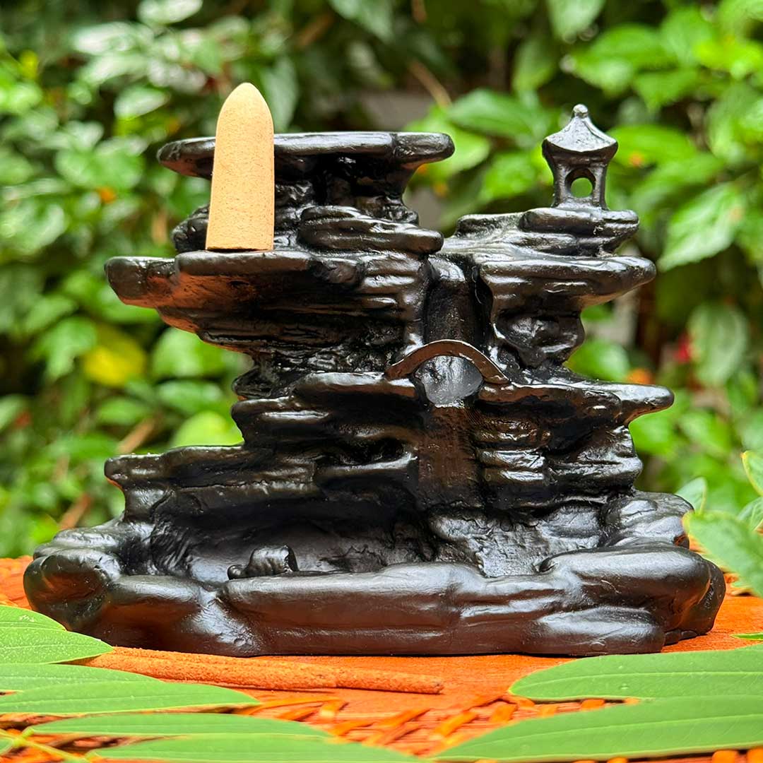 Incensário Cachoeira de Rozan Pelicano Azul em preto, com cascata esculpida e detalhes orientais, posicionado sobre superfície natural com folhas verdes.