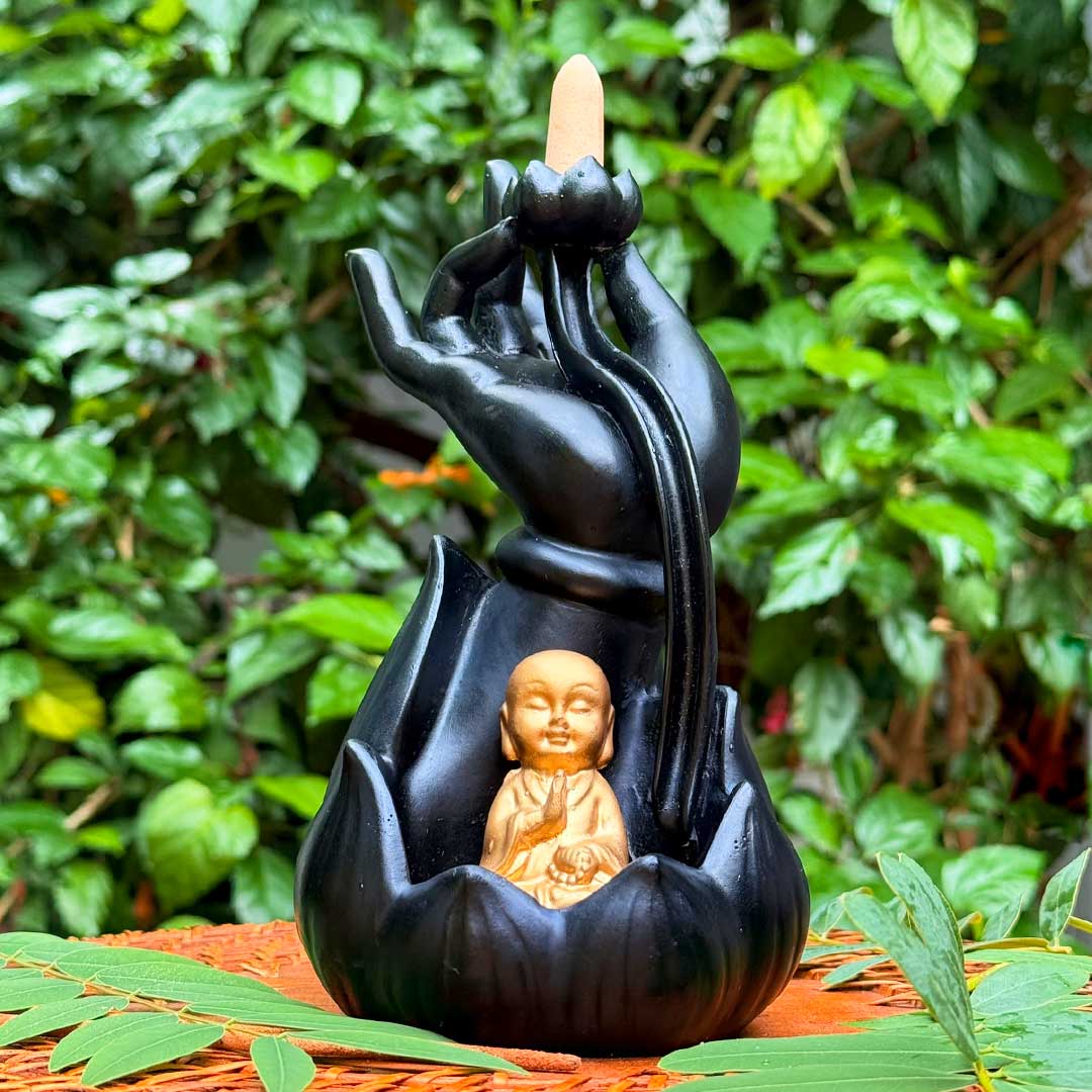 Incensário Mudra da Iluminação Pelicano Azul, com cone de incenso e uma estátua de Buda na base, cercado por folhagens verdes.