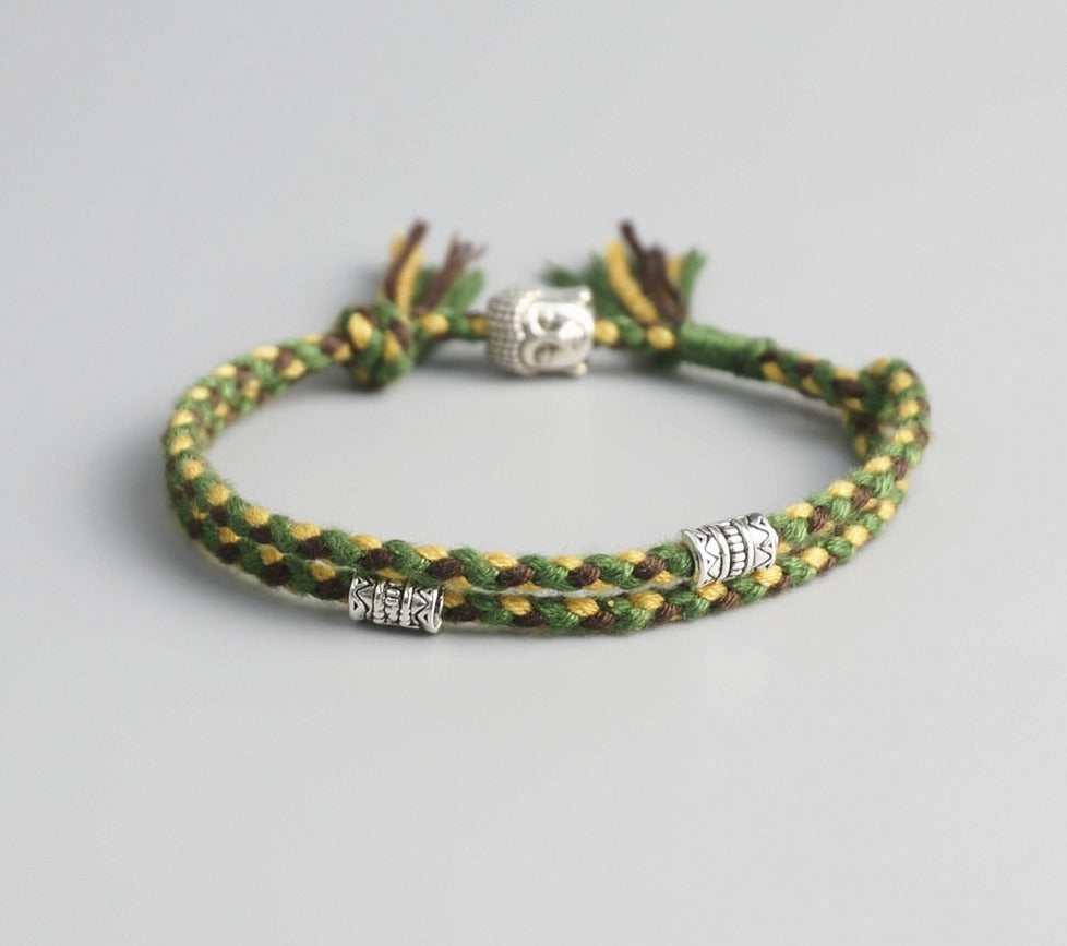 Pulseira Budista trançada em fios verde, amarelo e marrom, com detalhes prateados de Buda e símbolos espirituais. Pelicano Azul.
