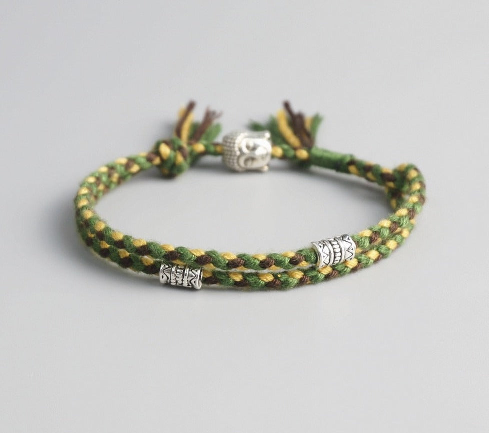 Pulseira Budista trançada em fios verdes e amarelos, com detalhes prateados de Buda e símbolos espirituais. Pelicano Azul.