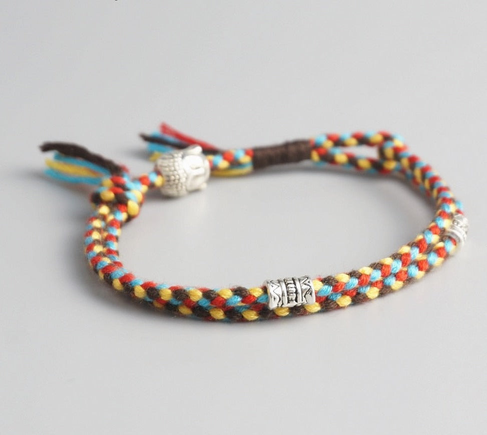 Pulseira Budista trançada com fios em vermelho, amarelo e azul, com detalhe de pingente prateado de Buda e contas decorativas. Pelicano Azul.