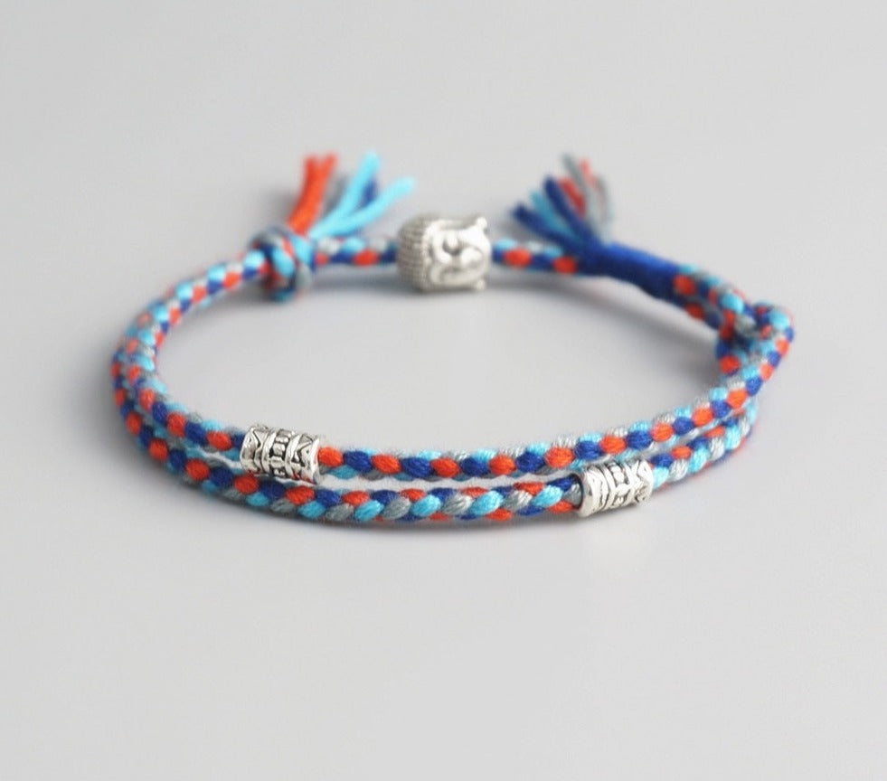 Pulseira Budista com fios trançados em azul, vermelho e laranja, com pingente de Buda e detalhes prateados. Pelicano Azul.
